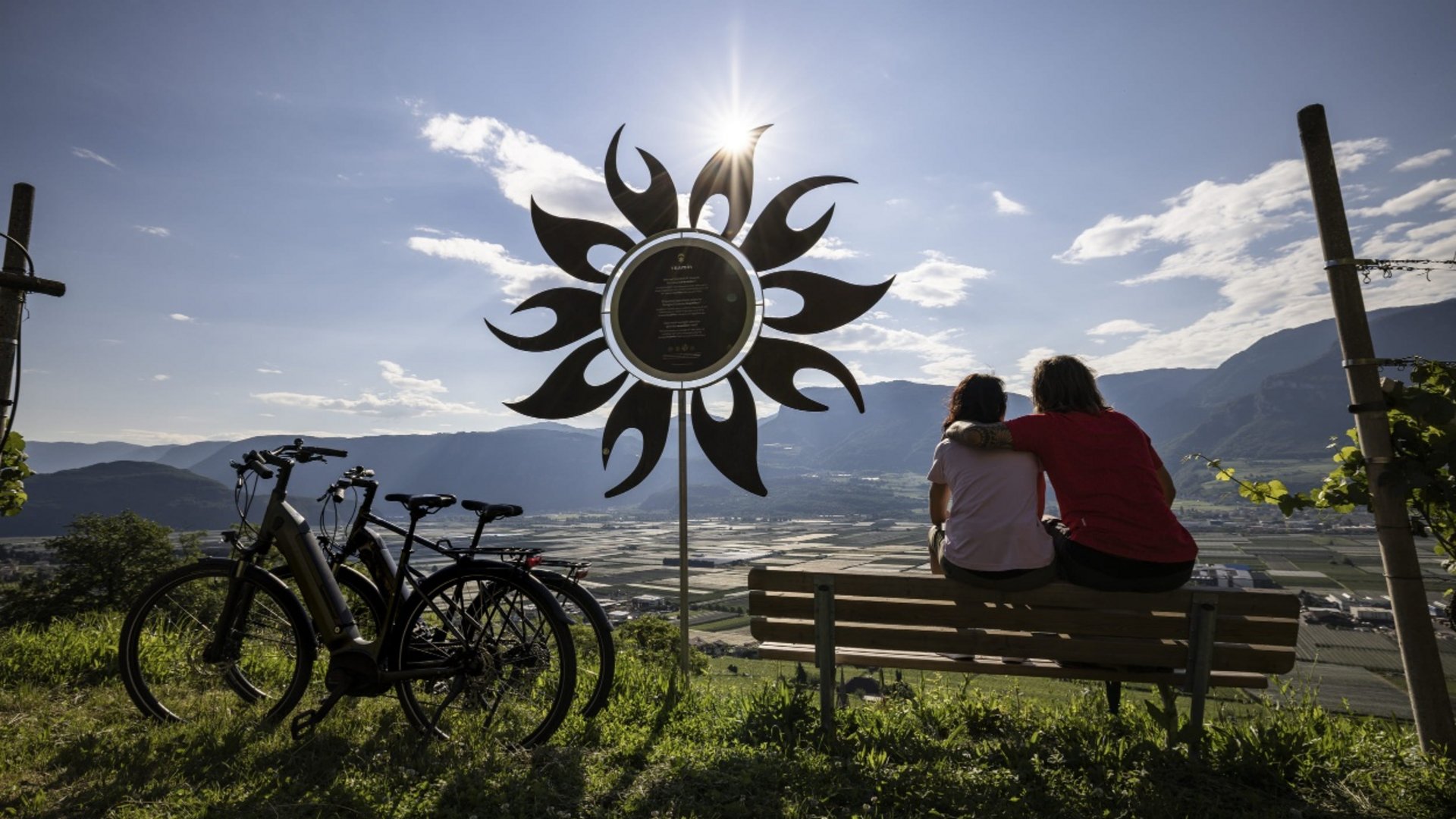 Couple sitting on bench by bikes with mountain valley and sun sculpture behind