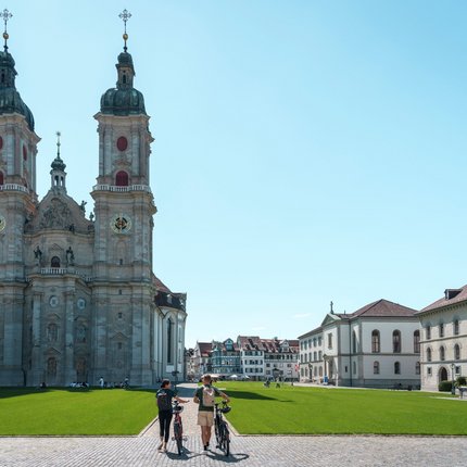 Two cyclists walking with bikes near the baroque St. Gallen Cathedral on a sunny day