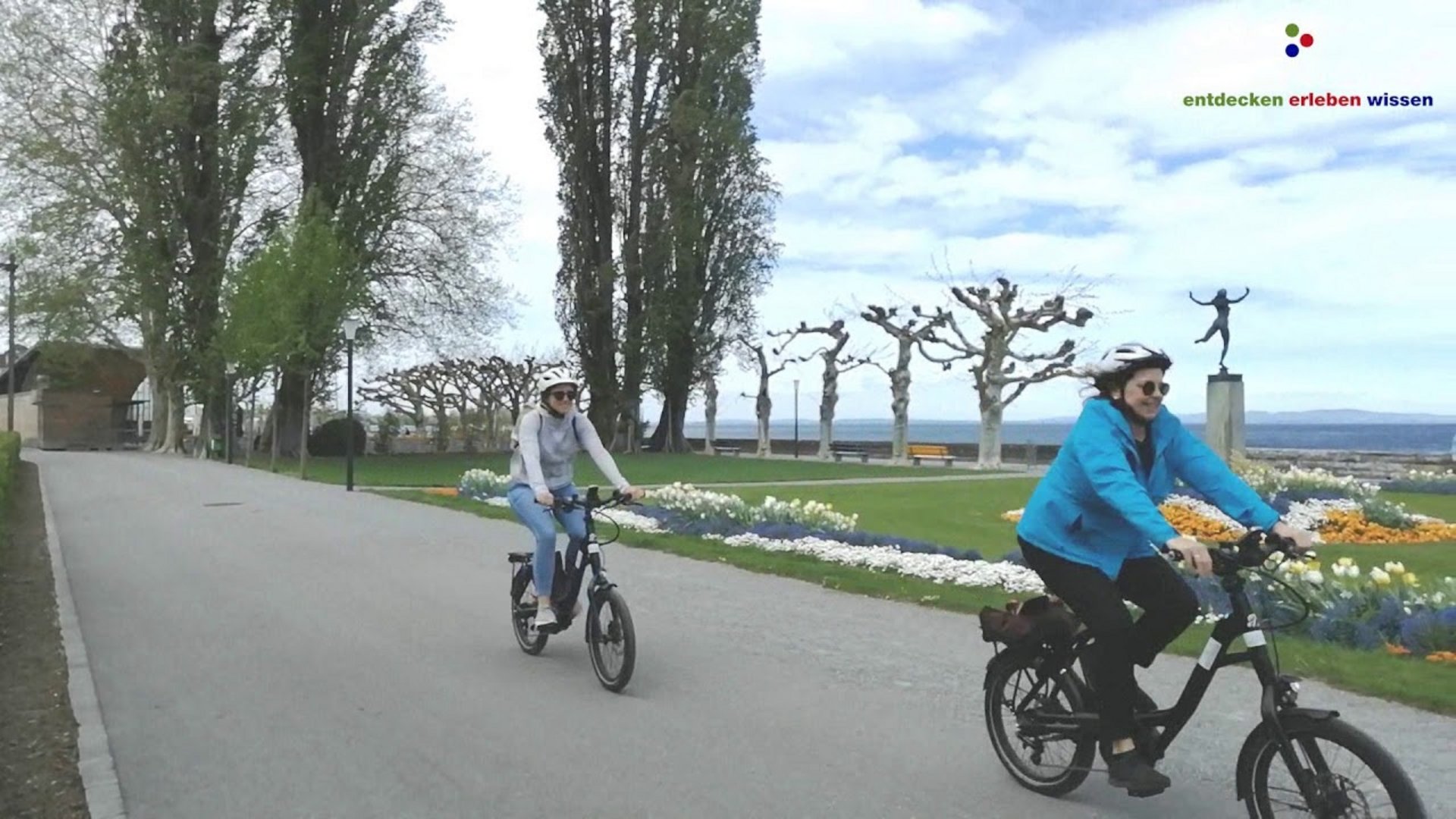Two people riding bicycles on a path next to flowerbeds and trees