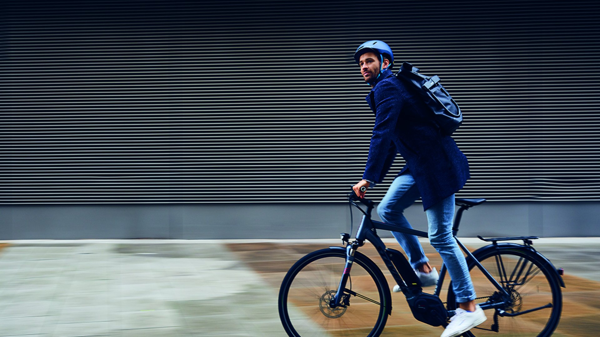Man wearing helmet and backpack riding a bicycle by a modern wall