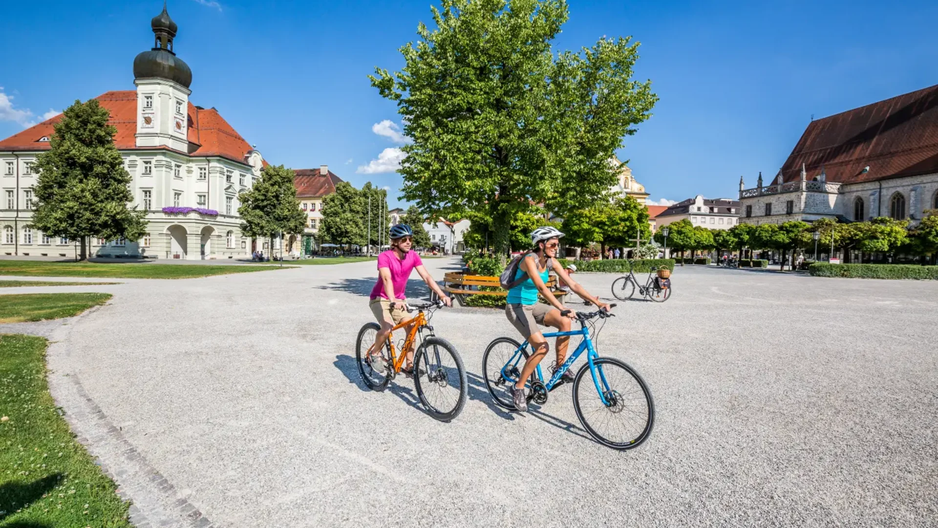 Zwei Radfahrer fahren auf einem Platz mit historischen Gebäuden bei klarem Himmel