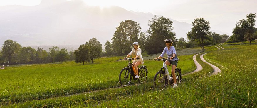Two women cycling on a grassy path under sunny sky