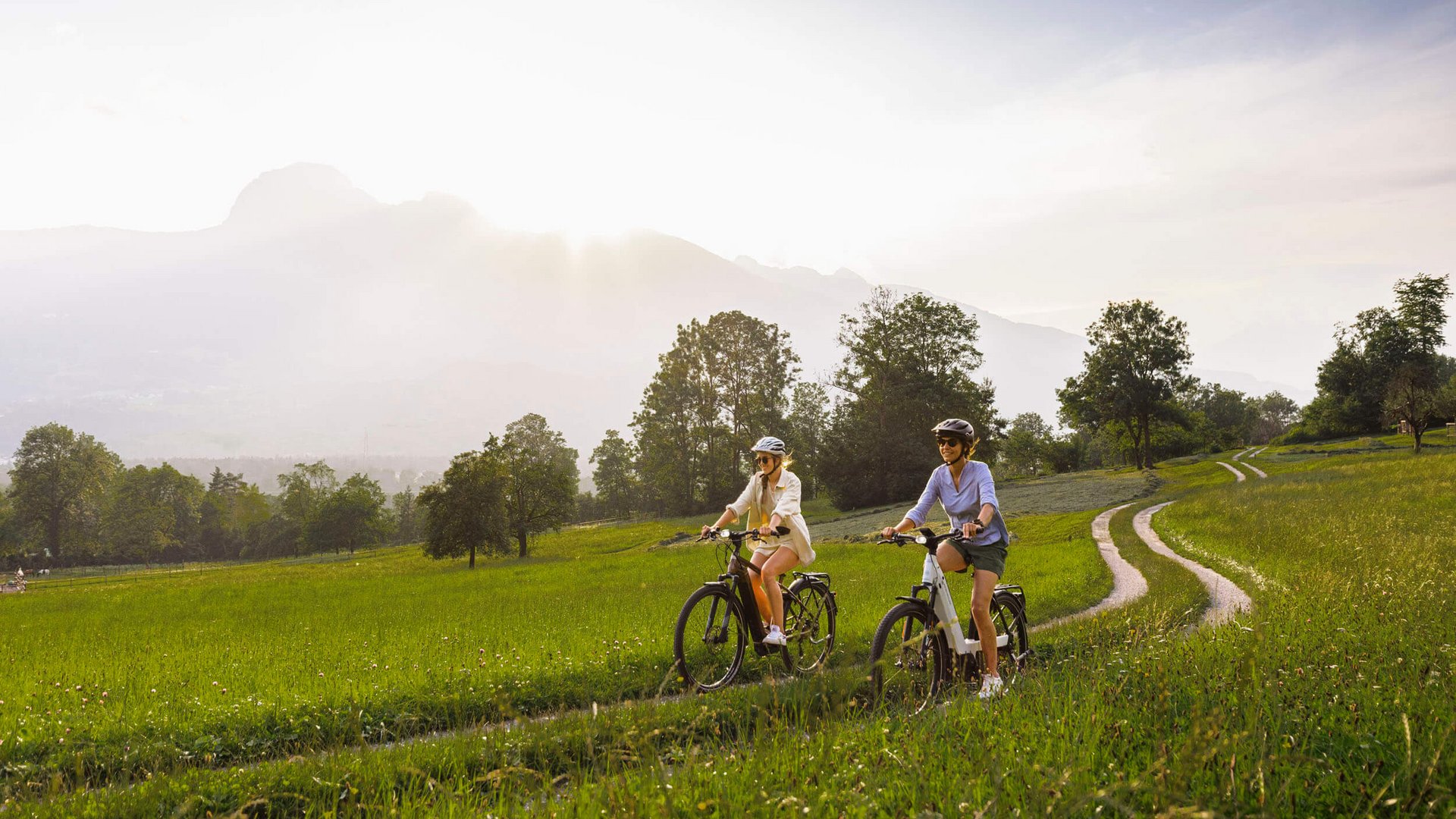 Two women cycling on a grassy path under sunny sky