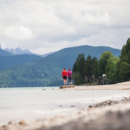 Zwei Radfahrer stehen an einem Seeufer mit Bergen und Wald im Hintergrund