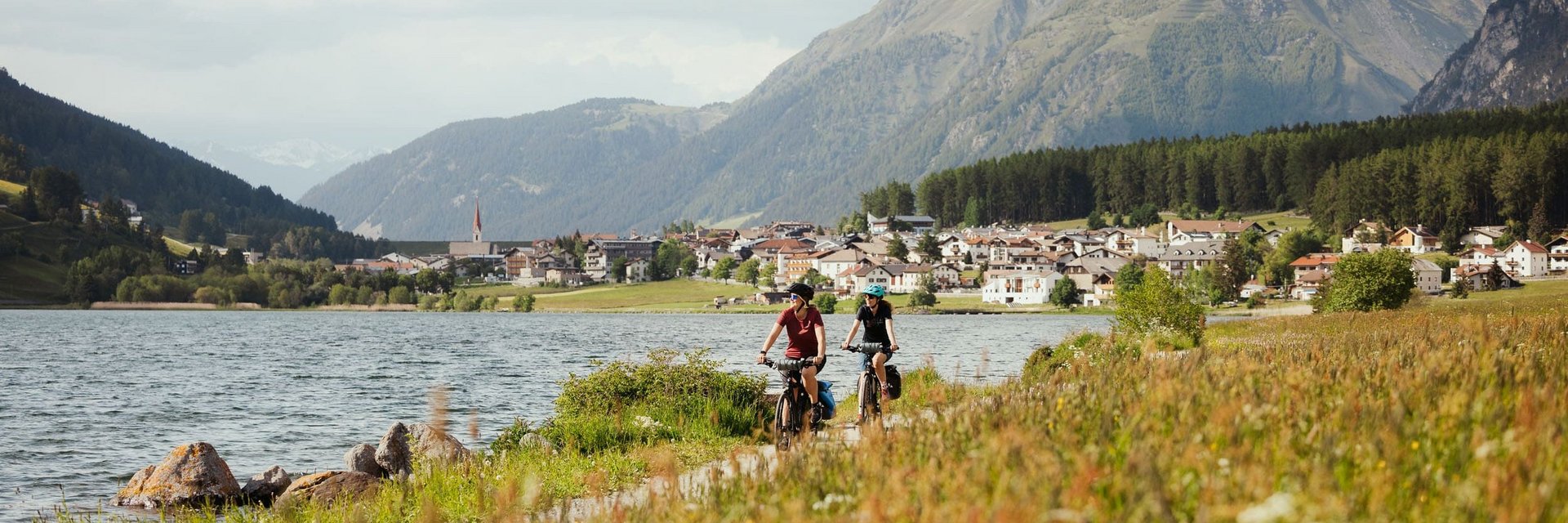Two cyclists riding by a lake with mountains and village in background