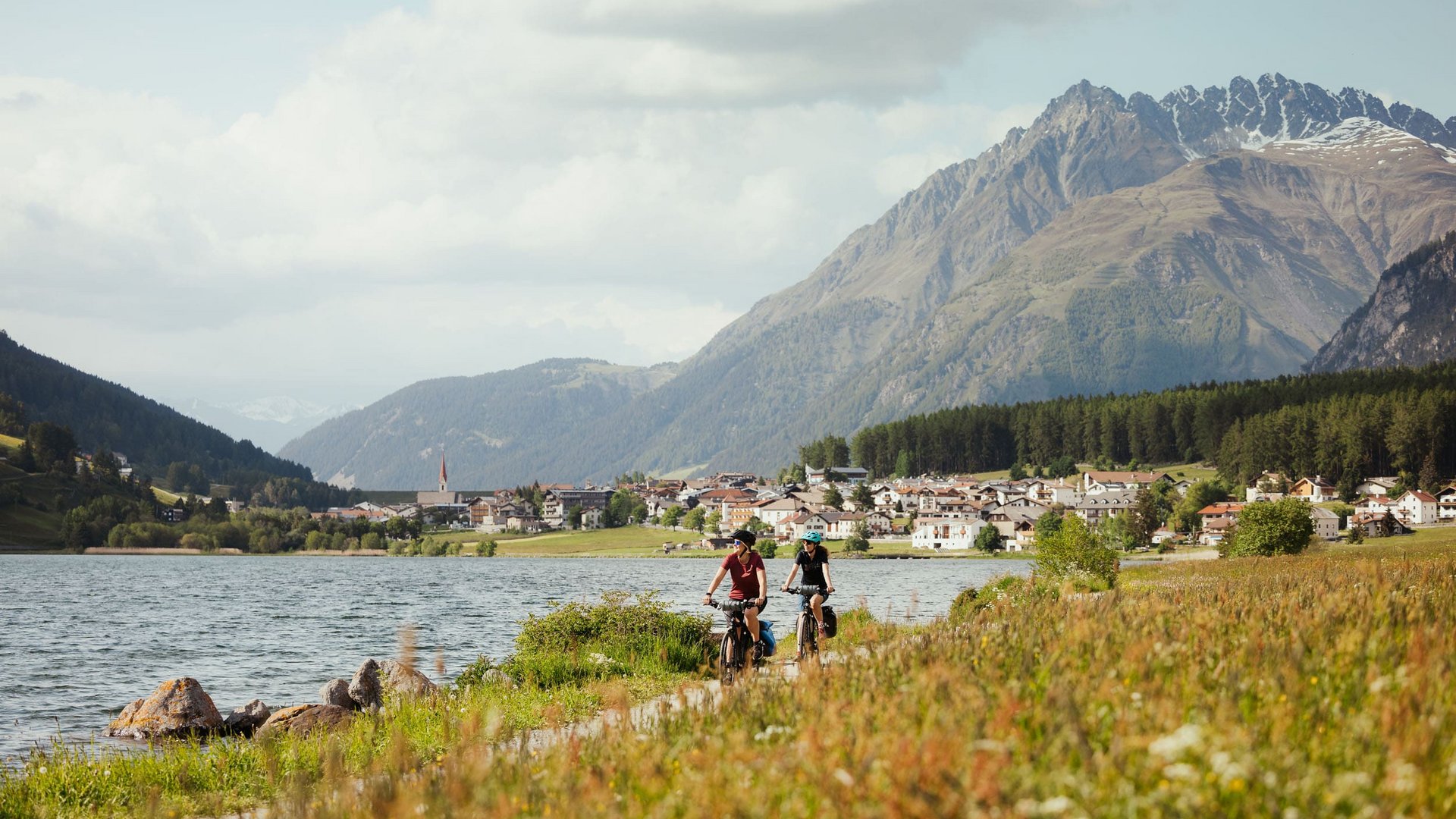 Two cyclists riding by a lake with mountains and village in background