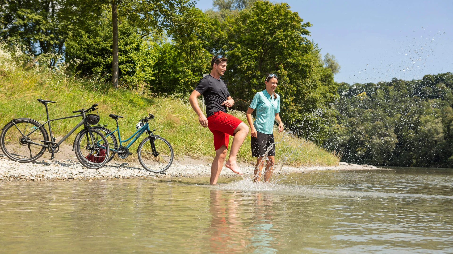 Zwei Menschen planschen mit Fahrrädern am Flussufer im Sommer