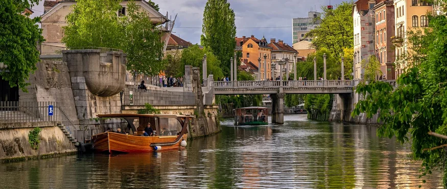 Flussfahrt mit Boot unter Brücke in einer Stadt mit historischen Gebäuden und Bäumen
