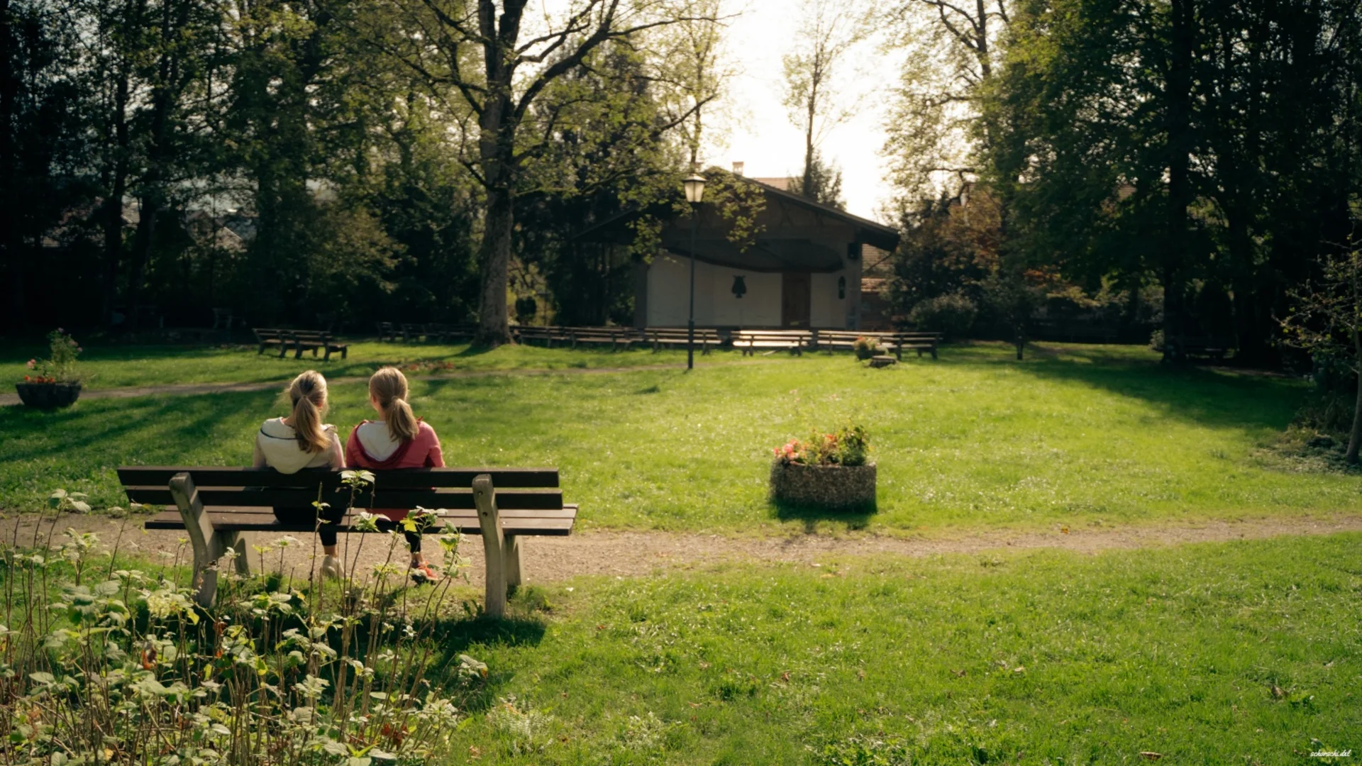 Zwei Mädchen sitzen auf einer Bank in einem sonnigen Park mit Bäumen und einem Haus