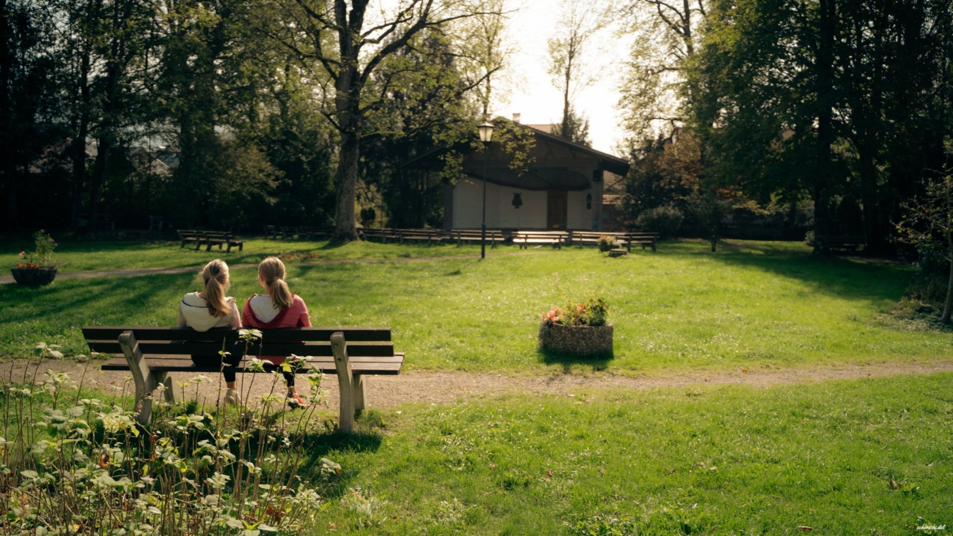 Two girls sitting on a bench in a sunny park with trees and a house