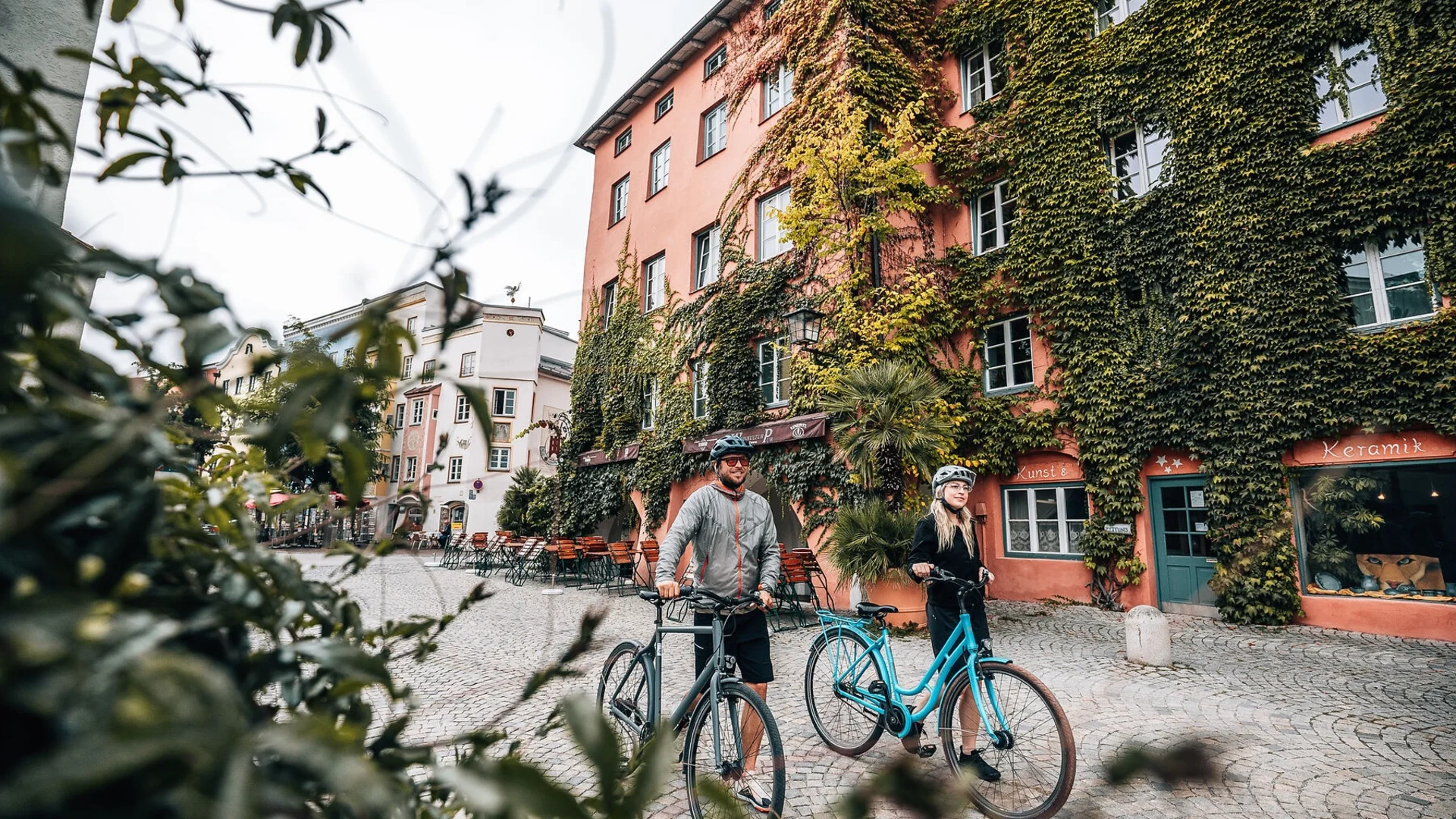 Zwei Personen mit Fahrrädern in einer Gasse mit Häusern, die mit Efeu bedeckt sind