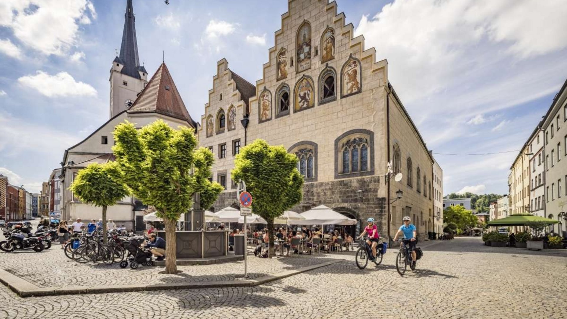 Busy square with cyclists, trees, and historic building on a sunny day