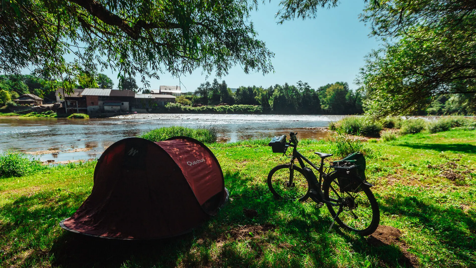 Tent and bicycle by the riverbank under trees on a sunny day