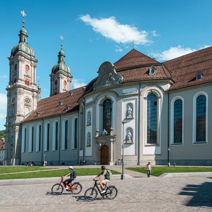 People cycling in front of a large historic church on a sunny day