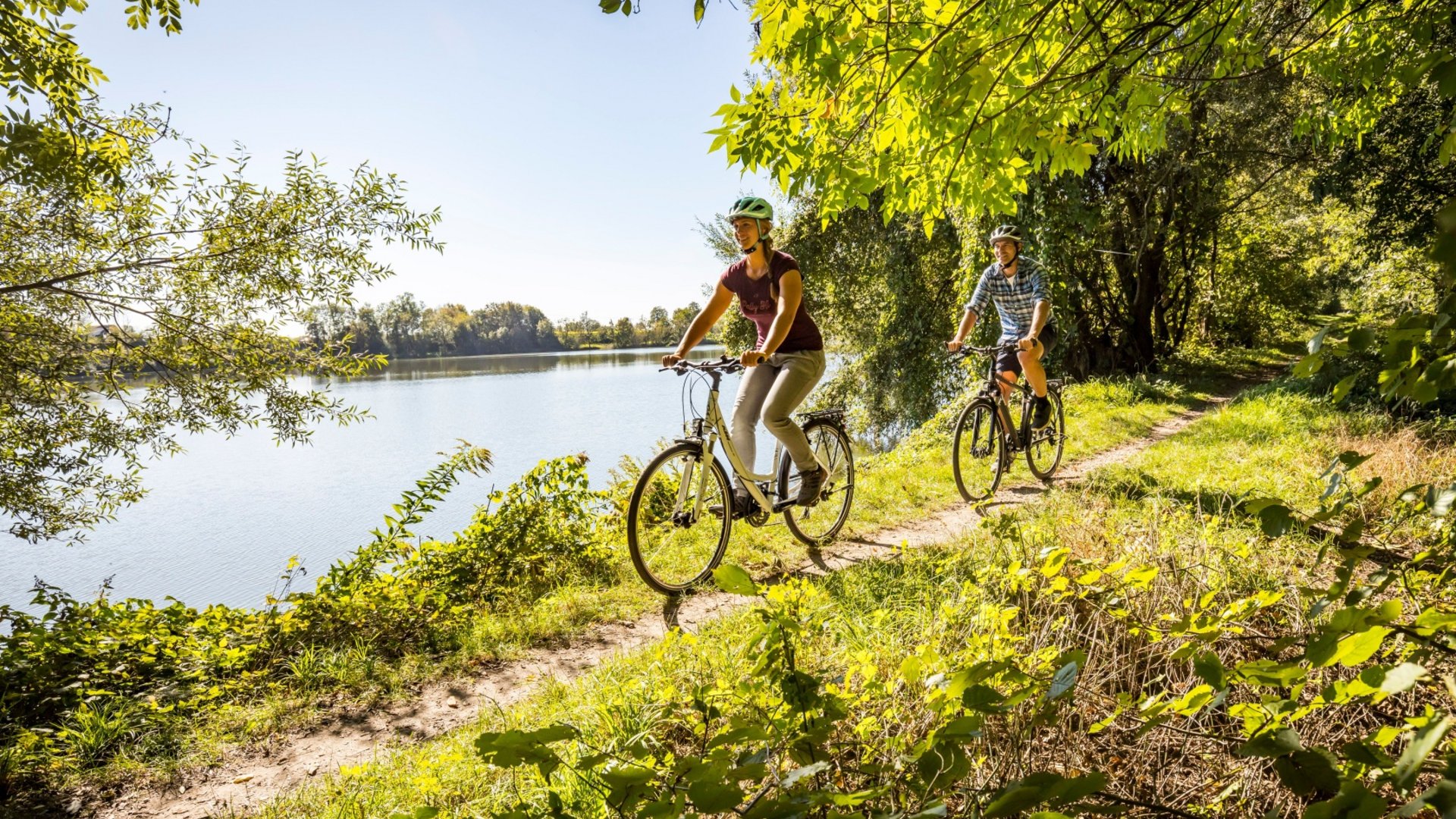 Two cyclists riding on a trail beside a river on a sunny day
