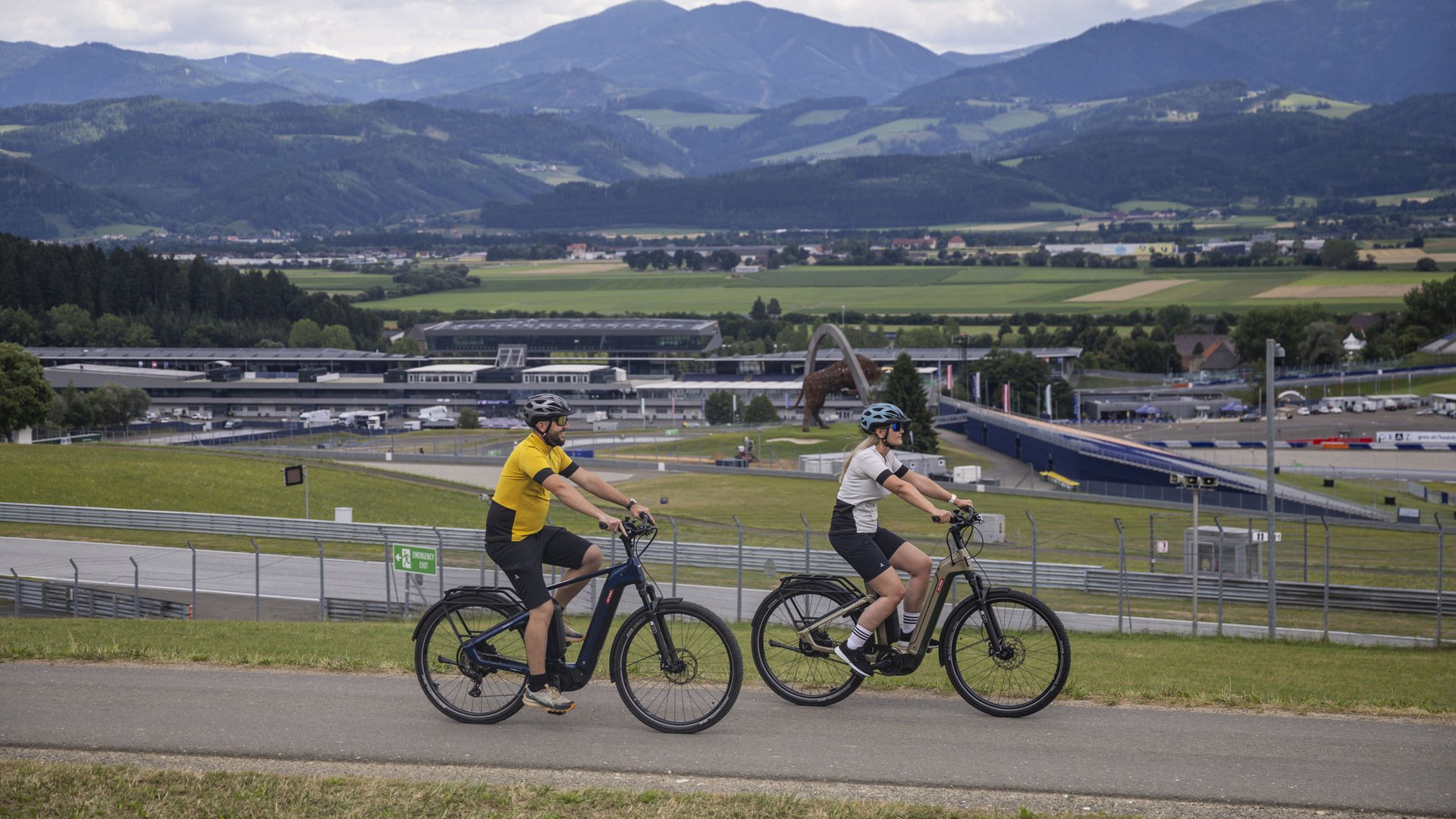 Two cyclists on e-bikes with mountains and racetrack in the background