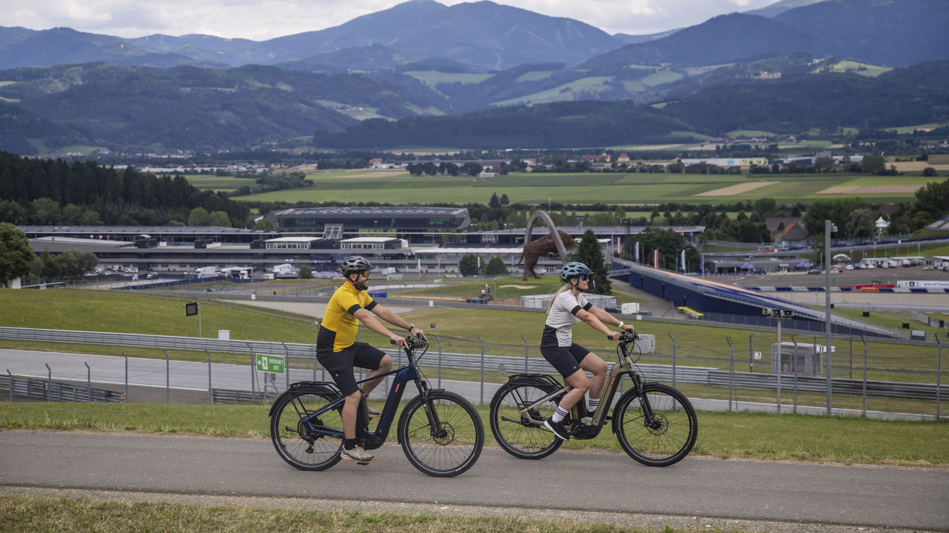 Twee fietsers op e-bikes met bergen en racecircuit op de achtergrond