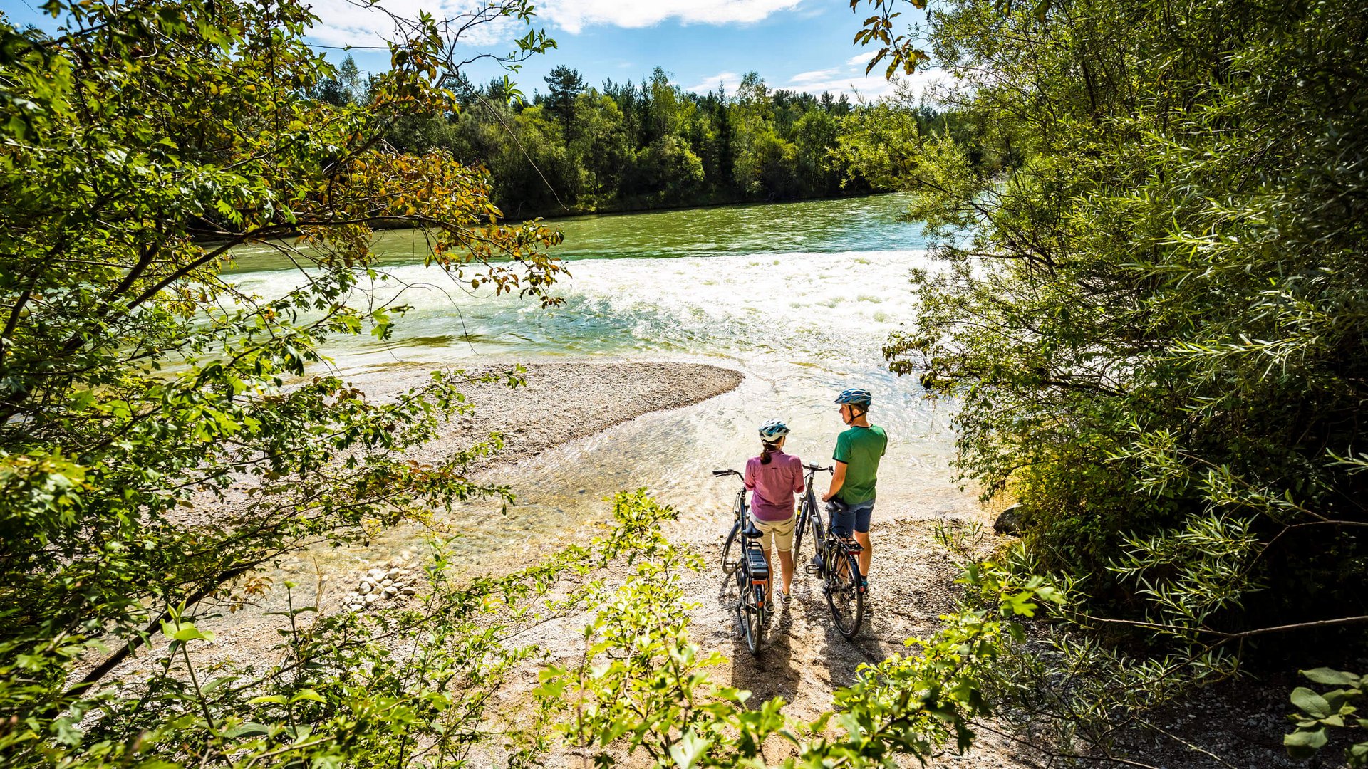 Two cyclists with helmets stand by the riverbank enjoying nature