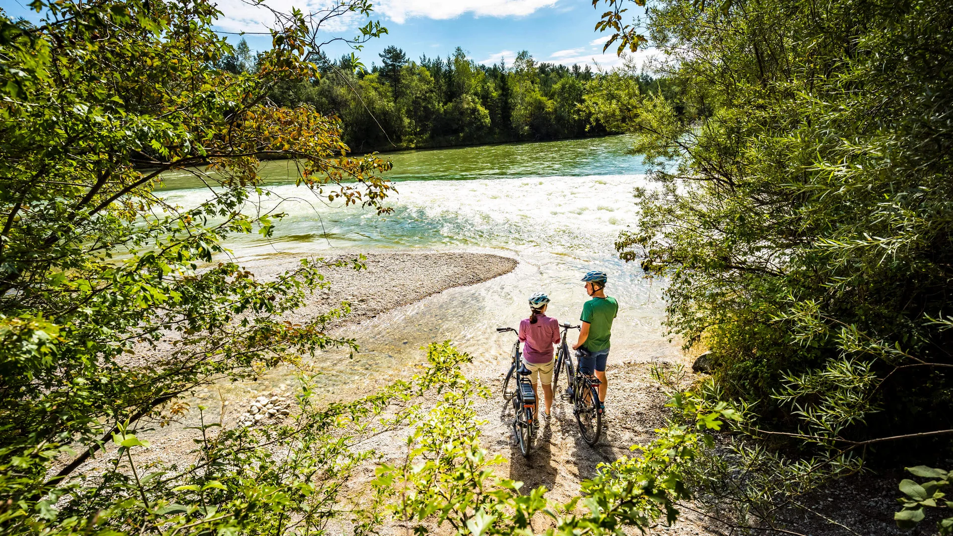 Zwei Radfahrer mit Helmen stehen am Flussufer und genießen die Natur