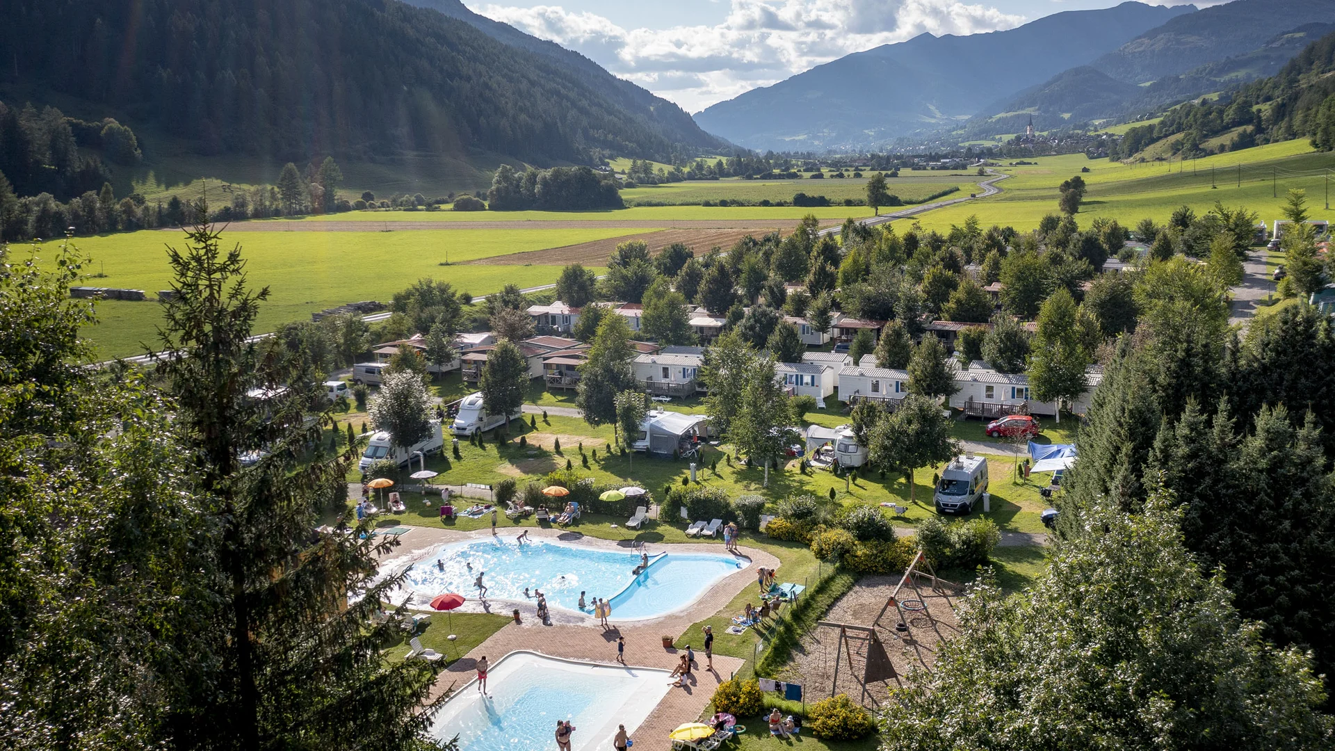 Camping in een dal met bergen, bomen en zwembaden onder een bewolkte lucht