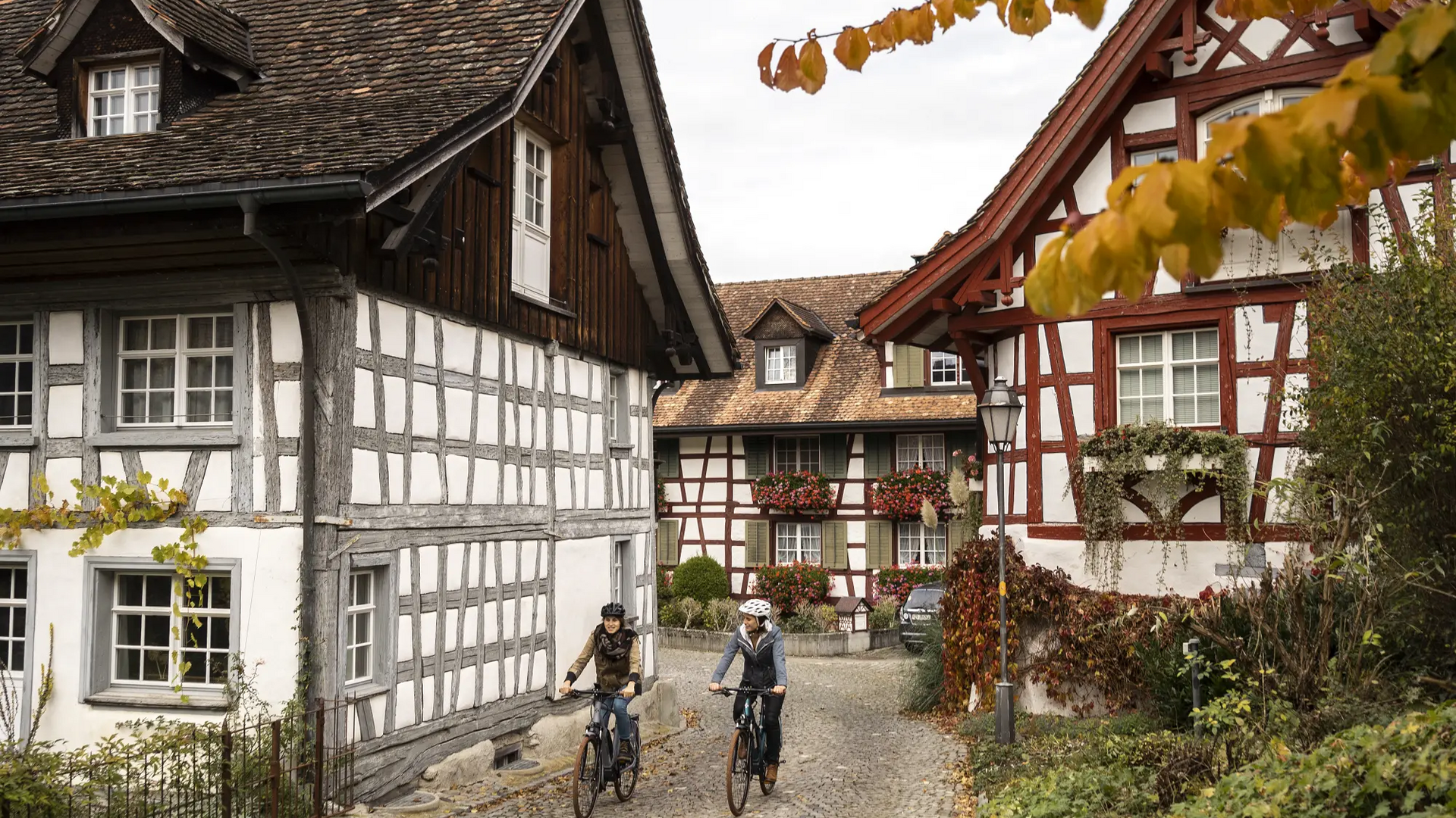 Two cyclists riding through a village with half-timbered houses in autumn