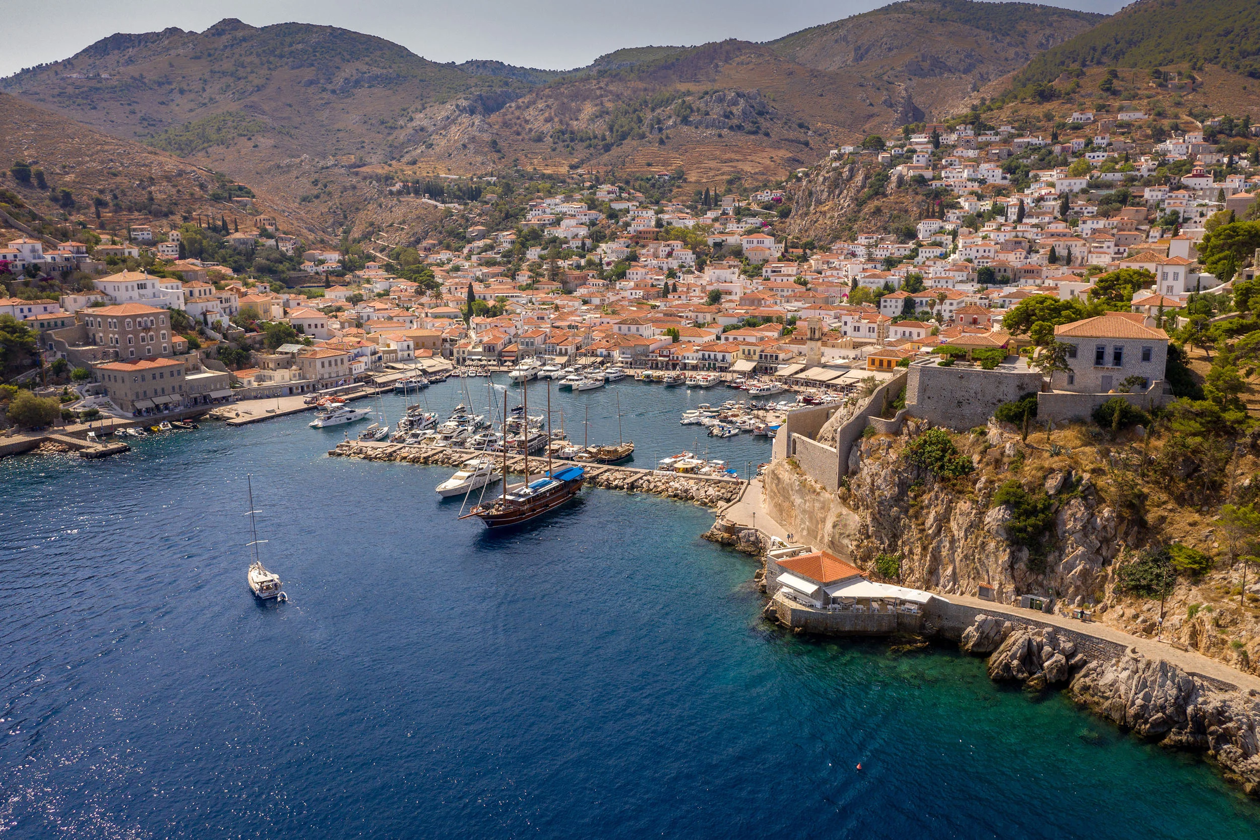 Luftaufnahme eines mediterranen Hafenorts mit Booten und Bergen im Hintergrund