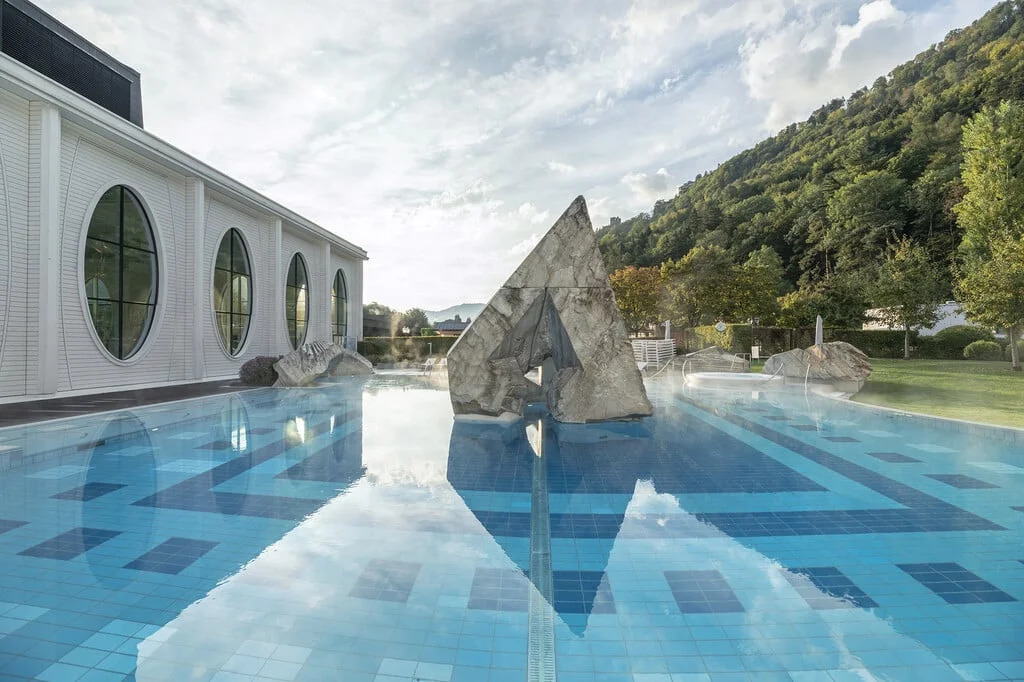 Außenpool mit geometrischer Marmor-Skulptur neben weißem Gebäude und Waldhügel