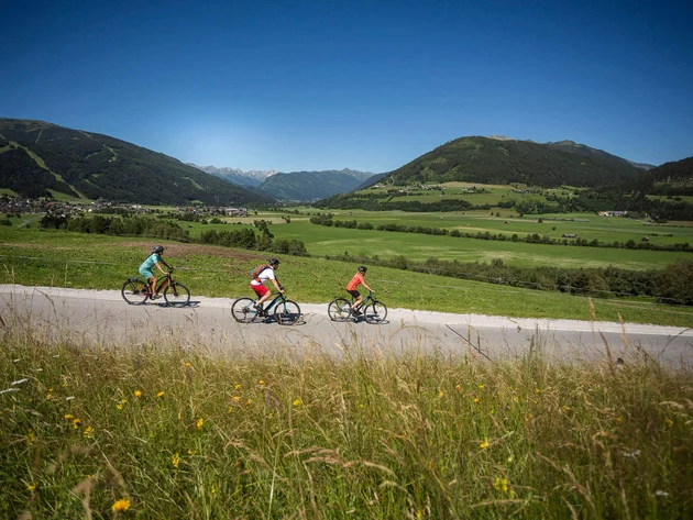 Drei Radfahrer auf Landstraße mit Alpenlandschaft im Hintergrund