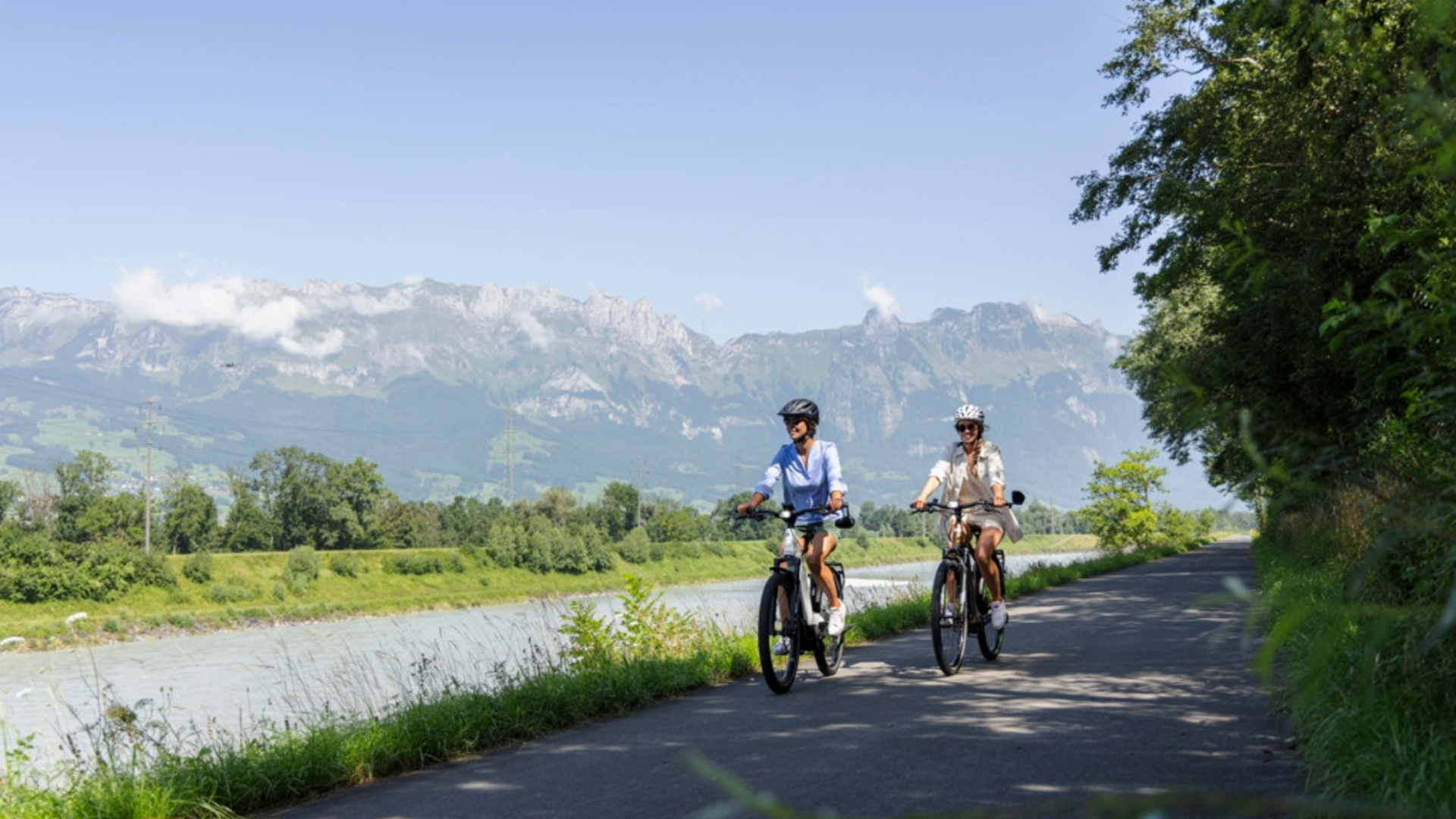 Two women cycling on a path beside a river with mountains in the background