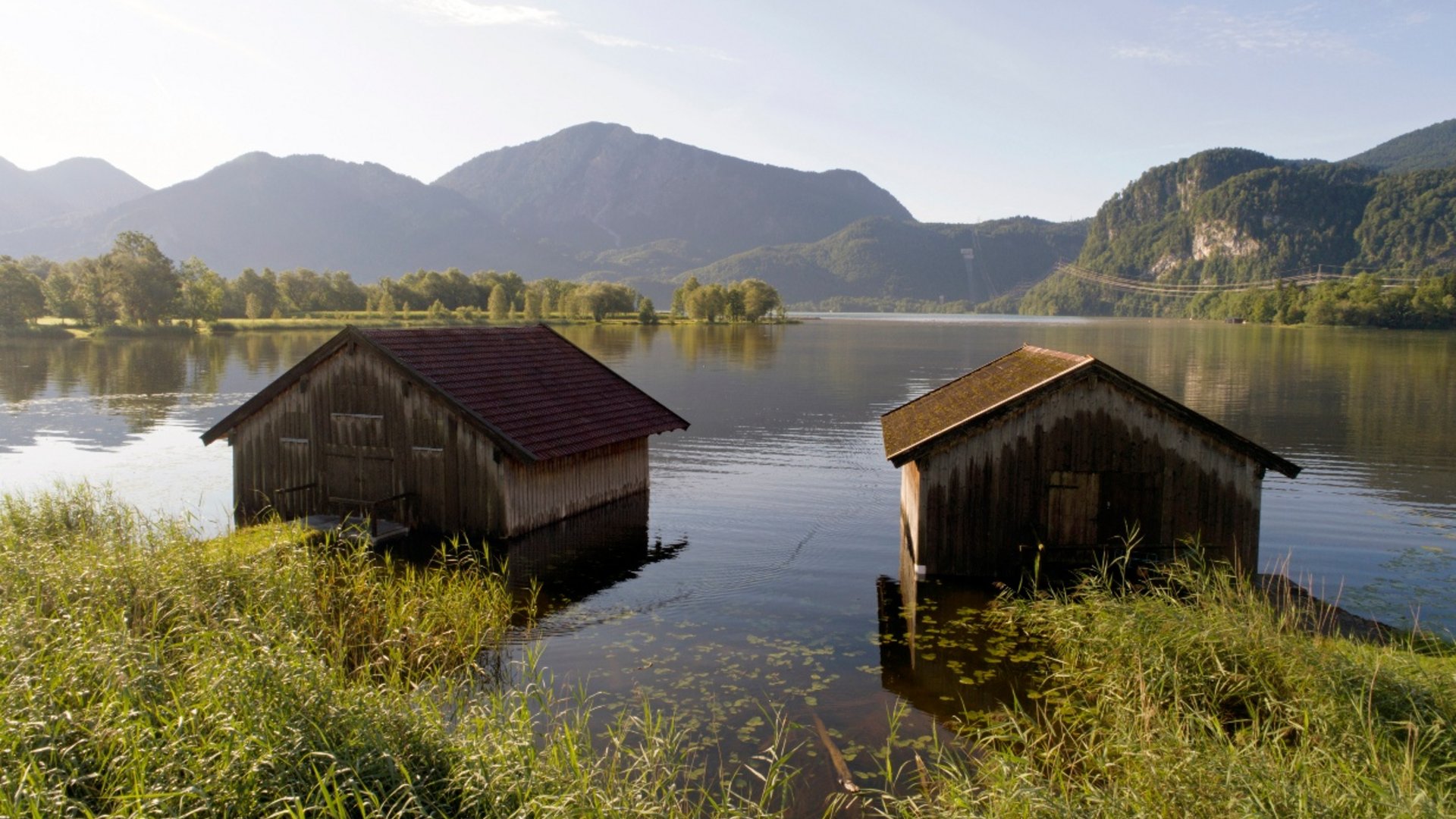 Two boathouses on a lake with mountains in the background in sunlight