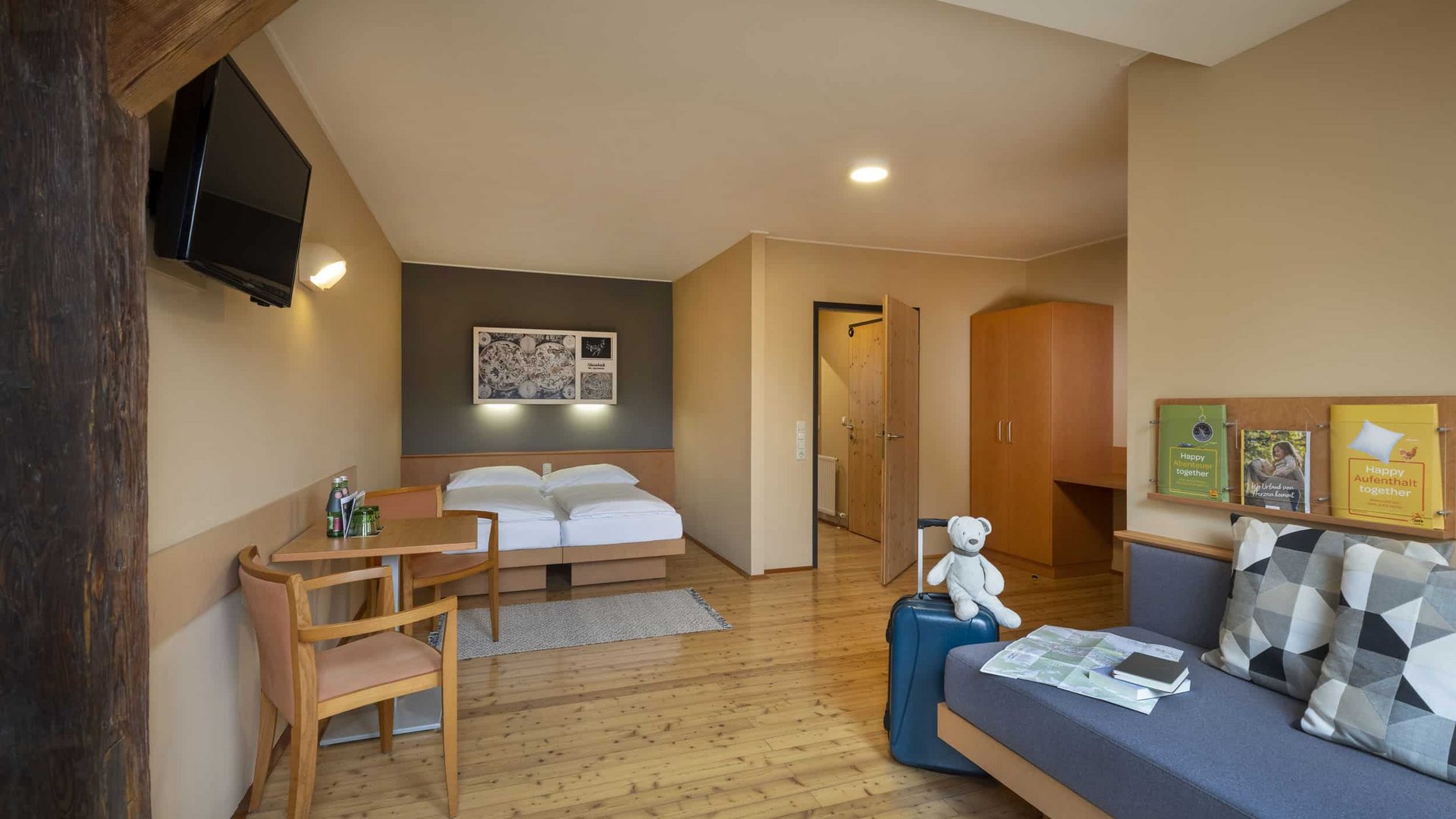 Cozy hotel room with double bed, table, sofa, and suitcase with teddy bear