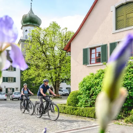 Schlössertour Region Frauenfeld © Balz Kubli Zwei Radfahrer fahren durch ein Dorf vor einer Kirche und Blumen