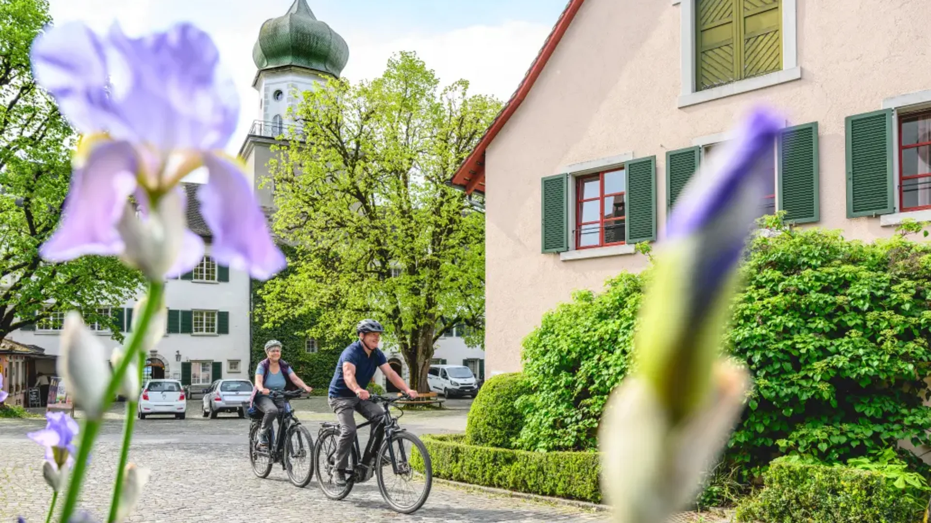 Schlössertour Region Frauenfeld © Balz Kubli Zwei Radfahrer fahren durch ein Dorf vor einer Kirche und Blumen