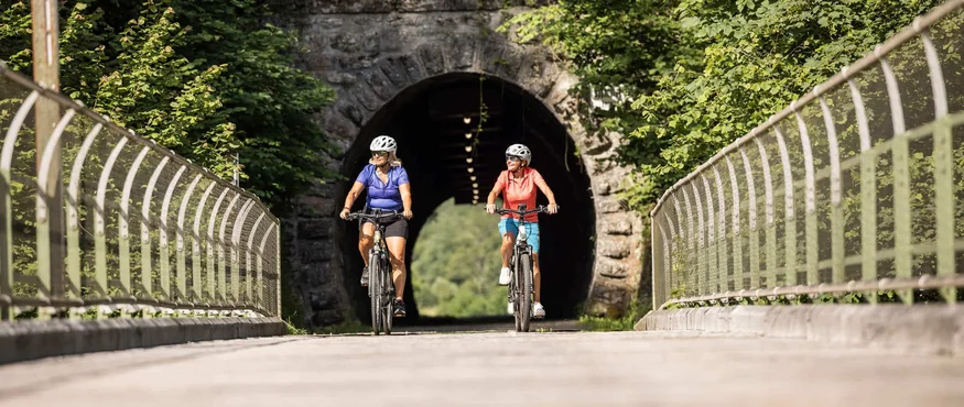 Twee vrouwen fietsen op een zonnig pad voor een tunnel omringd door groen