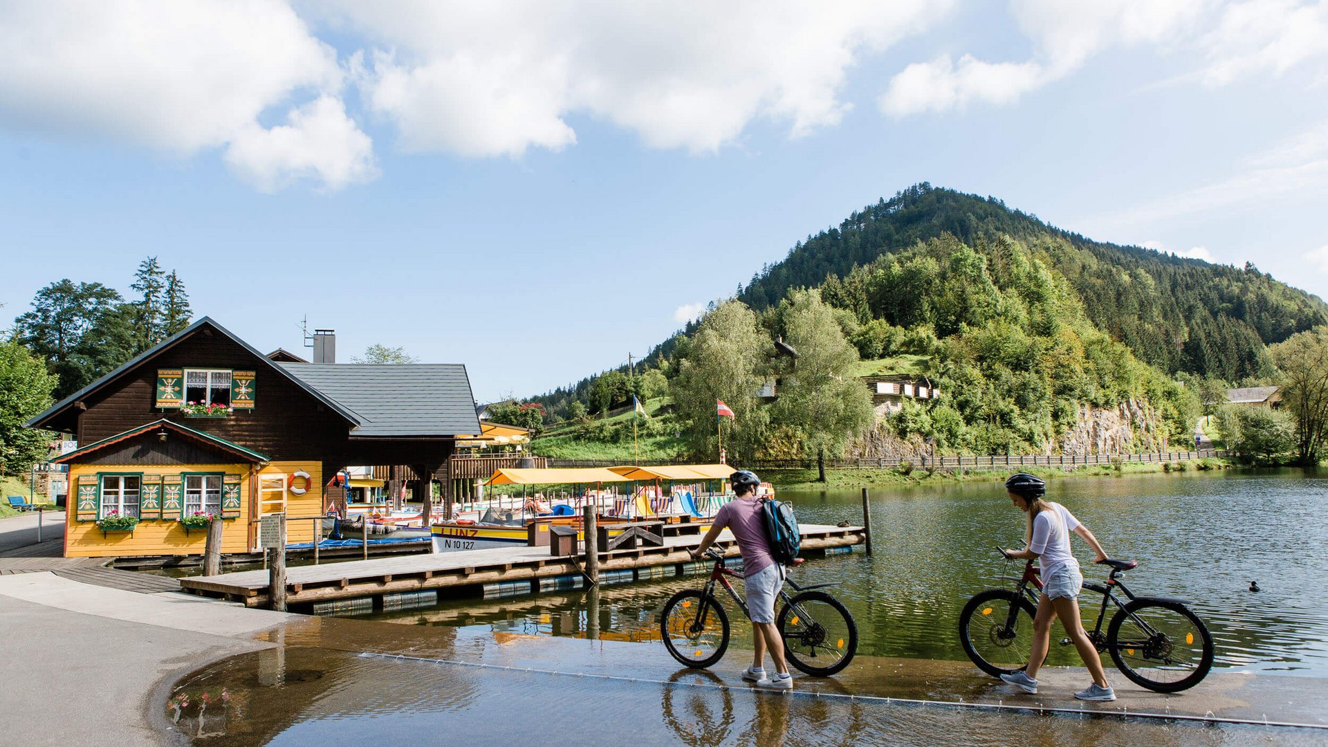 Two cyclists walking bikes beside lake with mountain and wooden house