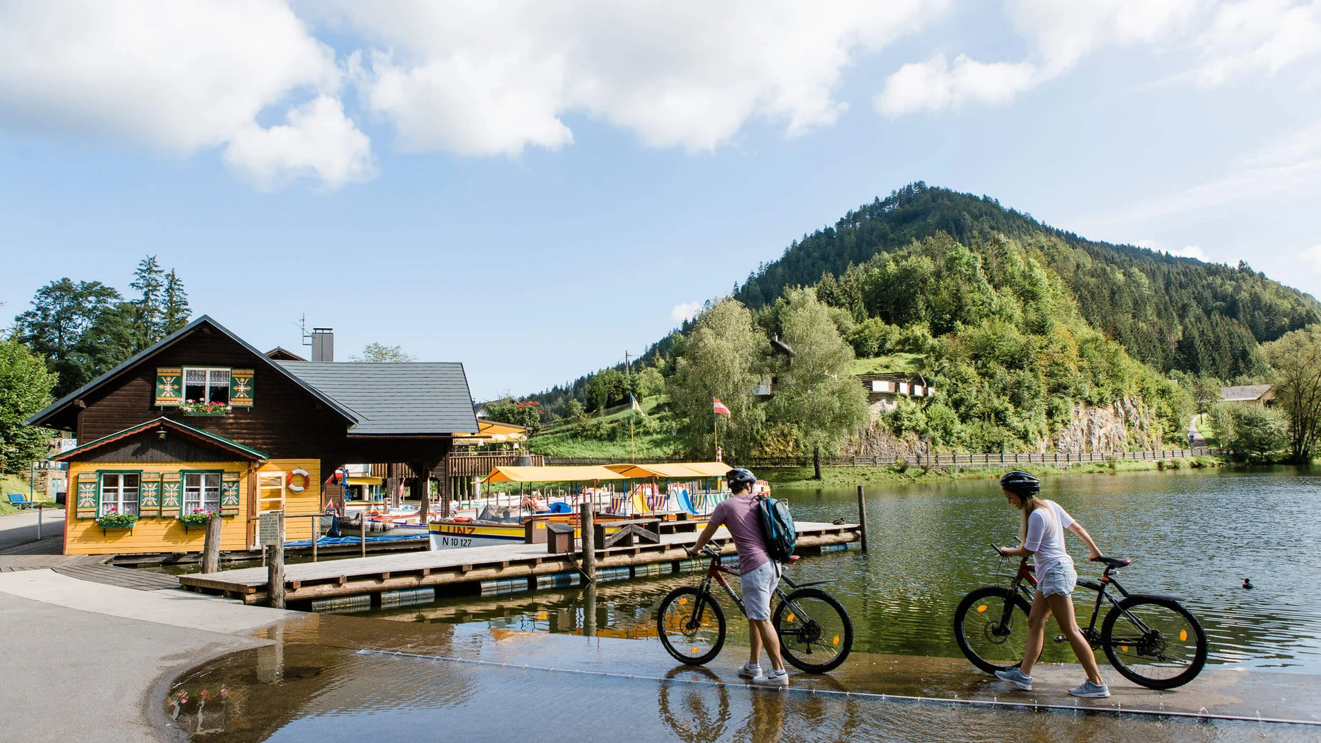 Zwei Radfahrer gehen mit ihren Fahrrädern an einem See mit Berg im Hintergrund