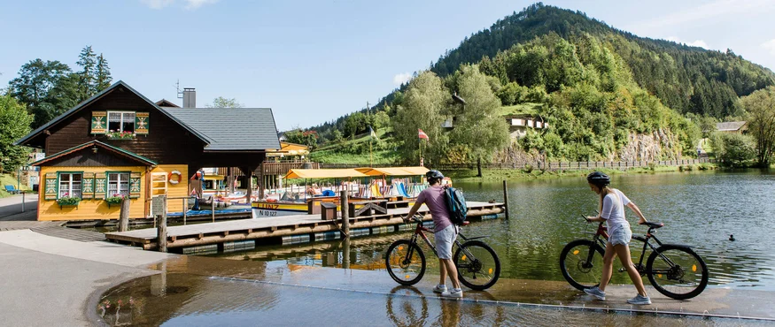 Zwei Radfahrer gehen mit ihren Fahrrädern an einem See mit Berg im Hintergrund
