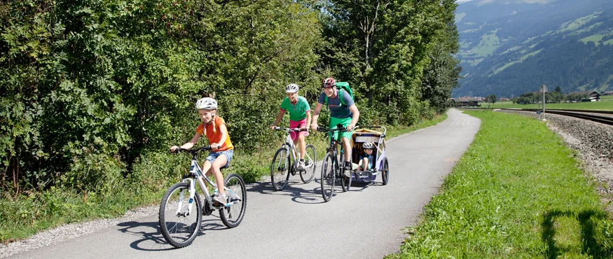 Familie fährt mit Fahrrädern auf einem Weg neben Bäumen und Bergen