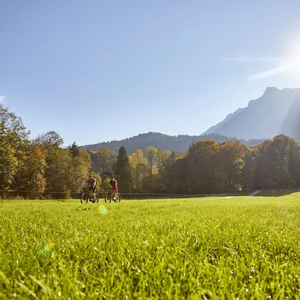 Zwei Radfahrer auf grüner Wiese mit Bergen und Sonne im Hintergrund