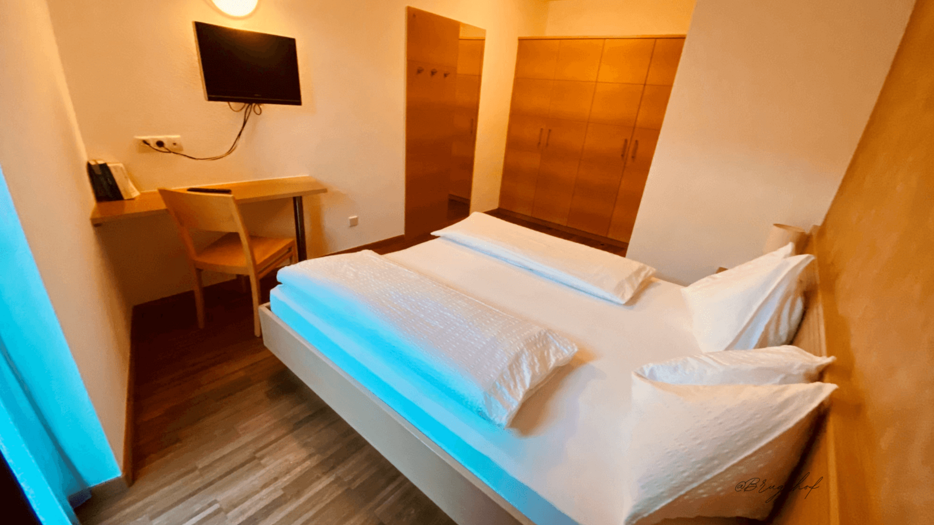 Small, simply furnished hotel room with bed, desk, and wall-mounted TV