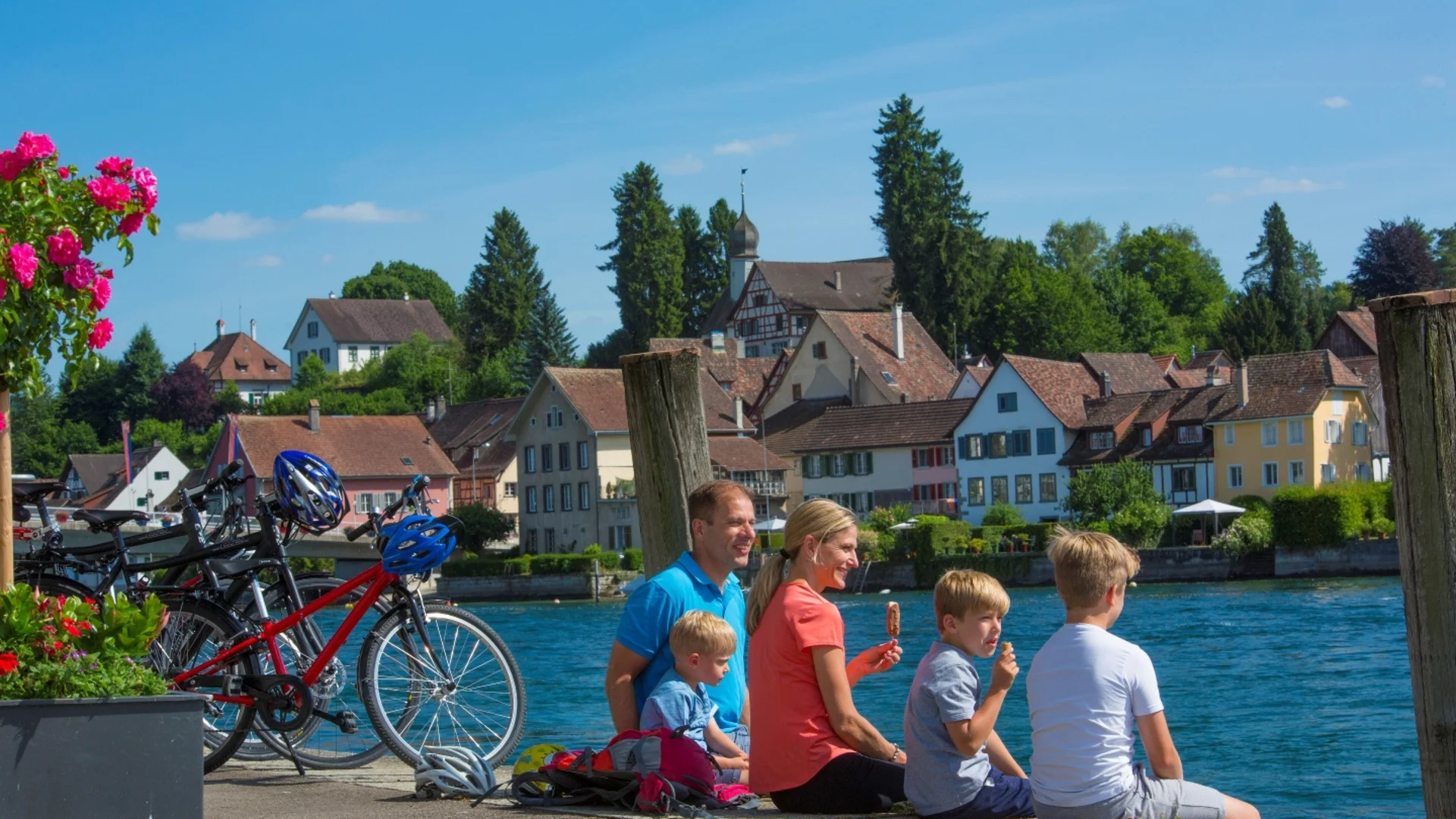 Familie zit aan rivier met fietsen bij huizen op een zonnige dag