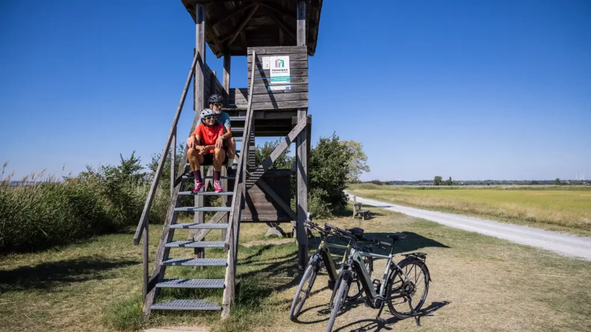 Zwei Radfahrer sitzen auf einer Holzbeobachtungsplattform neben ihren Fahrrädern auf einem Feldweg