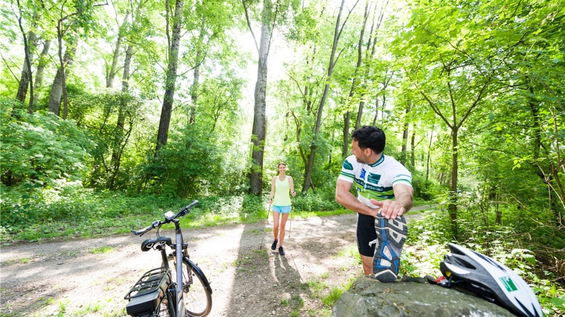 Man stretching in forest path as woman approaches with Nordic walking poles