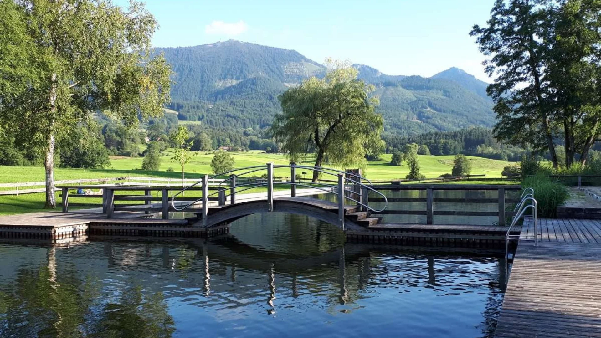 Holzbrücke über Wasser mit grünen Wiesen und Bergen im Hintergrund