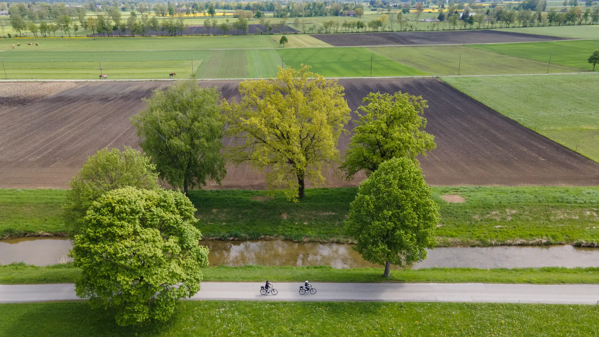Two cyclists on road beside river with farmland and mountains in background