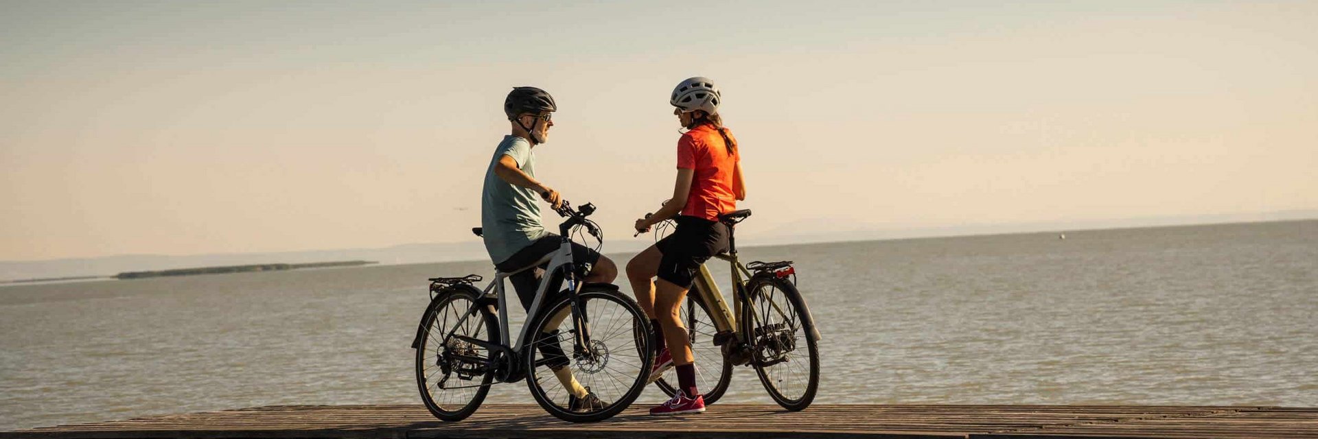 Two cyclists standing with bicycles on a pier by the water at sunset