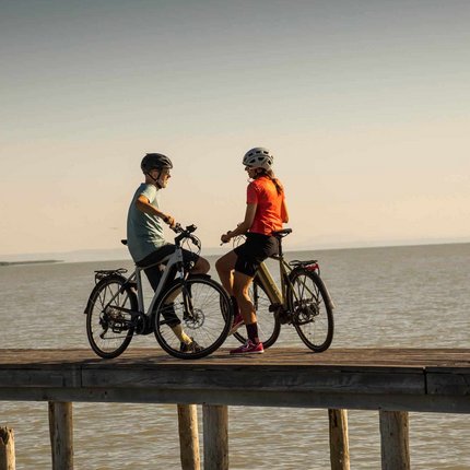 Two cyclists standing with bicycles on a pier by the water at sunset