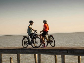 Two cyclists standing with bicycles on a pier by the water at sunset
