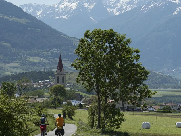 Ein Genuss-Radtag am Reschensee © IDM Südtirol - T. Grüner Zwei Radfahrer auf Landstraße vor Dorf und verschneiten Bergen