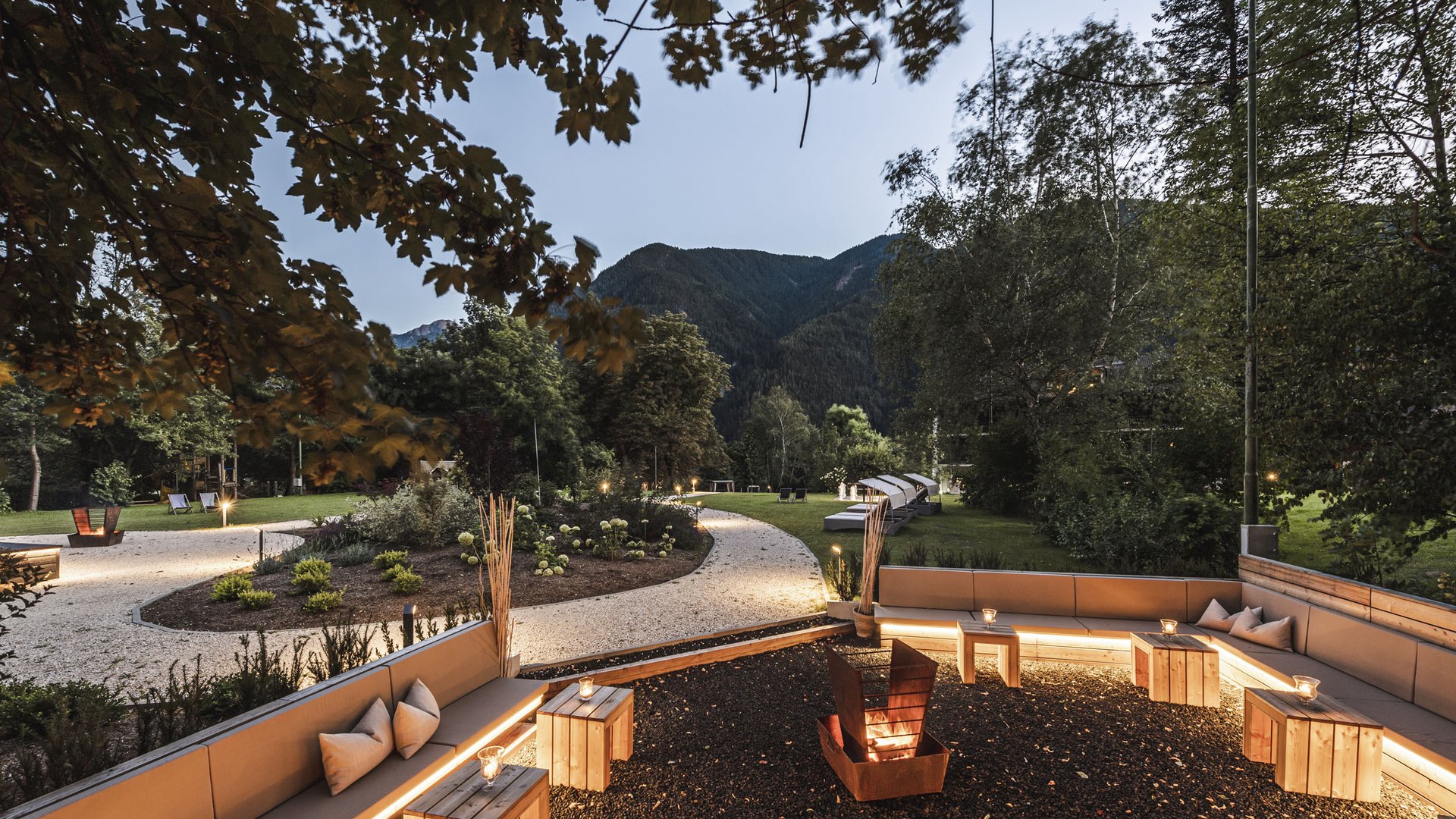 Lit garden with seating area and fire pit in front of mountain backdrop at dusk